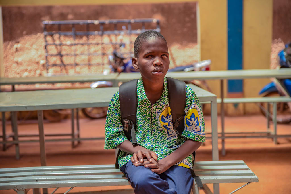 Kader is wearing a blue, white, and green patterned shirt, blue pants, and a black back pack. He is sitting outside his school on a green bench.